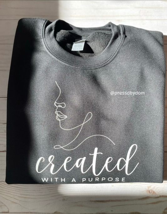 Folded black sweatshirt with white minimalist line-art face and text reading “Created With a Purpose” – faith-based apparel by Press’d by Dom.